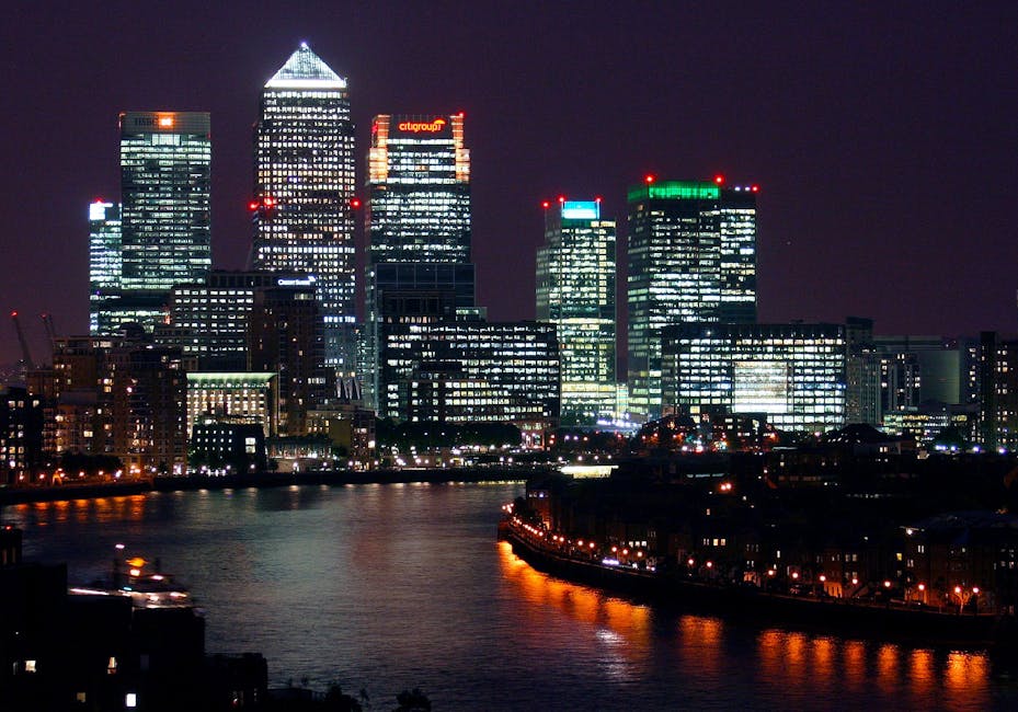 Nighttime cityscape of Canary Wharf with illuminated modern glass skyscrapers, including the One Canada Square building, reflecting on the river below. The tall buildings feature bright interior light