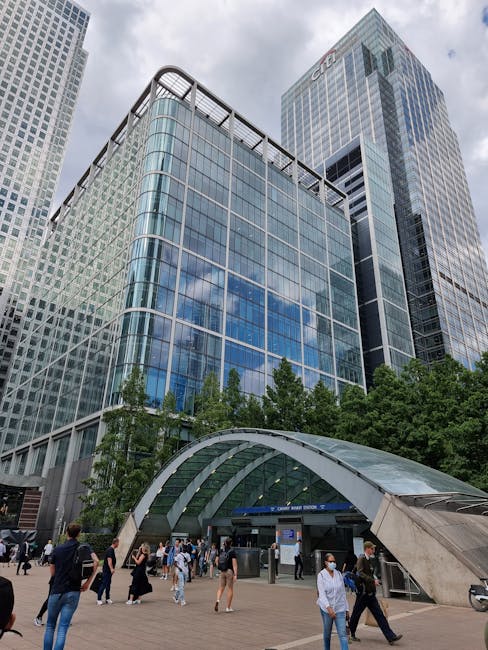 Nighttime cityscape of Canary Wharf with illuminated modern glass skyscrapers, including the One Canada Square building, reflecting on the river below. The tall buildings feature bright interior light