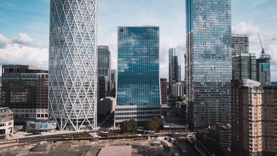 A high-angle view of a modern city skyline at sunset, featuring tall glass skyscrapers with reflective surfaces, detailed with visible windows and structural lines, set against a backdrop of a colourf