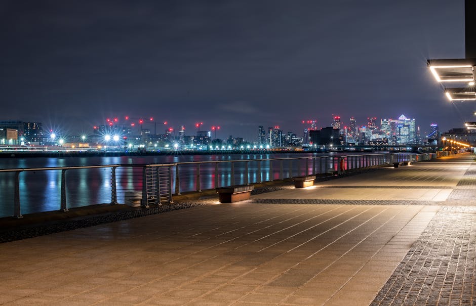 A panoramic view of West India Quay in Canary Wharf, featuring a marina with several moored boats and yachts, including a large wooden vessel and smaller sailboats, alongside a walkway with pedestrian